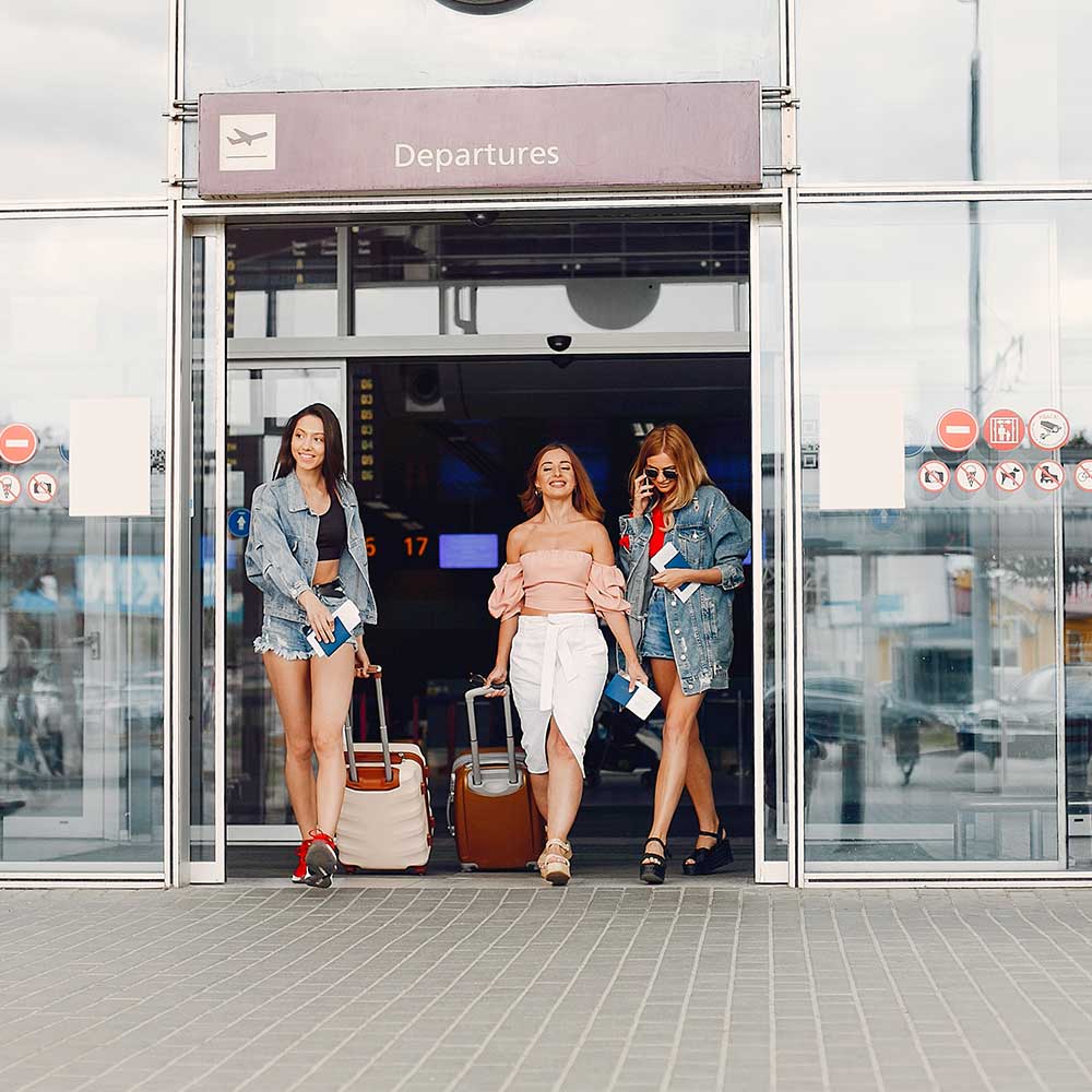 Three happy young women exiting the airport with their luggage, ready to start their trip